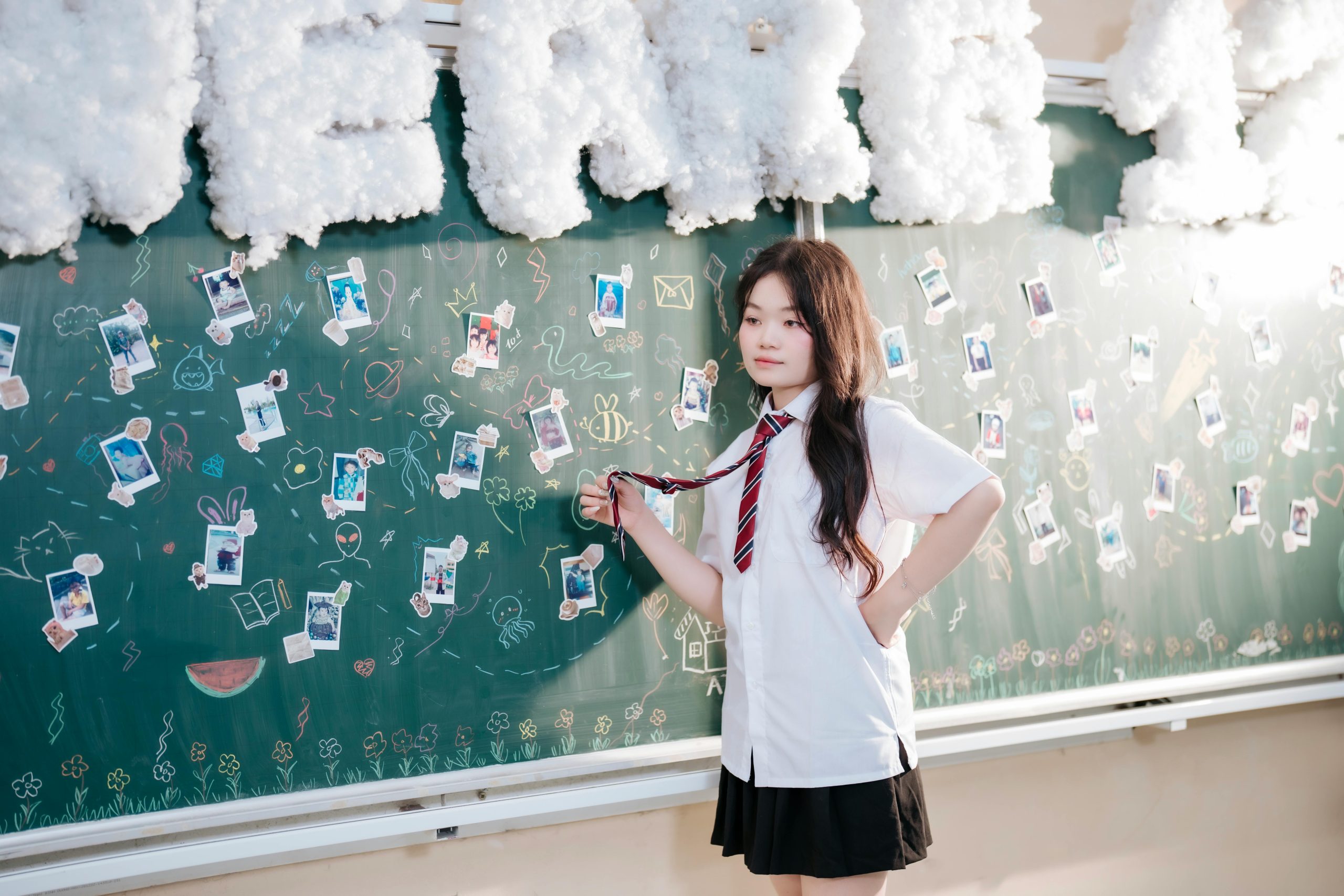 A young girl in a school uniform stands confidently by a creatively decorated chalkboard in a classroom.