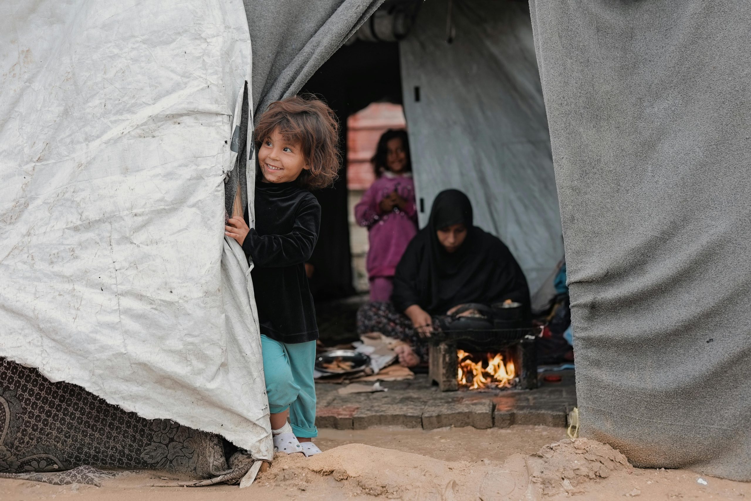 A young girl joyfully peering out of a tent in Gaza, with family members inside.