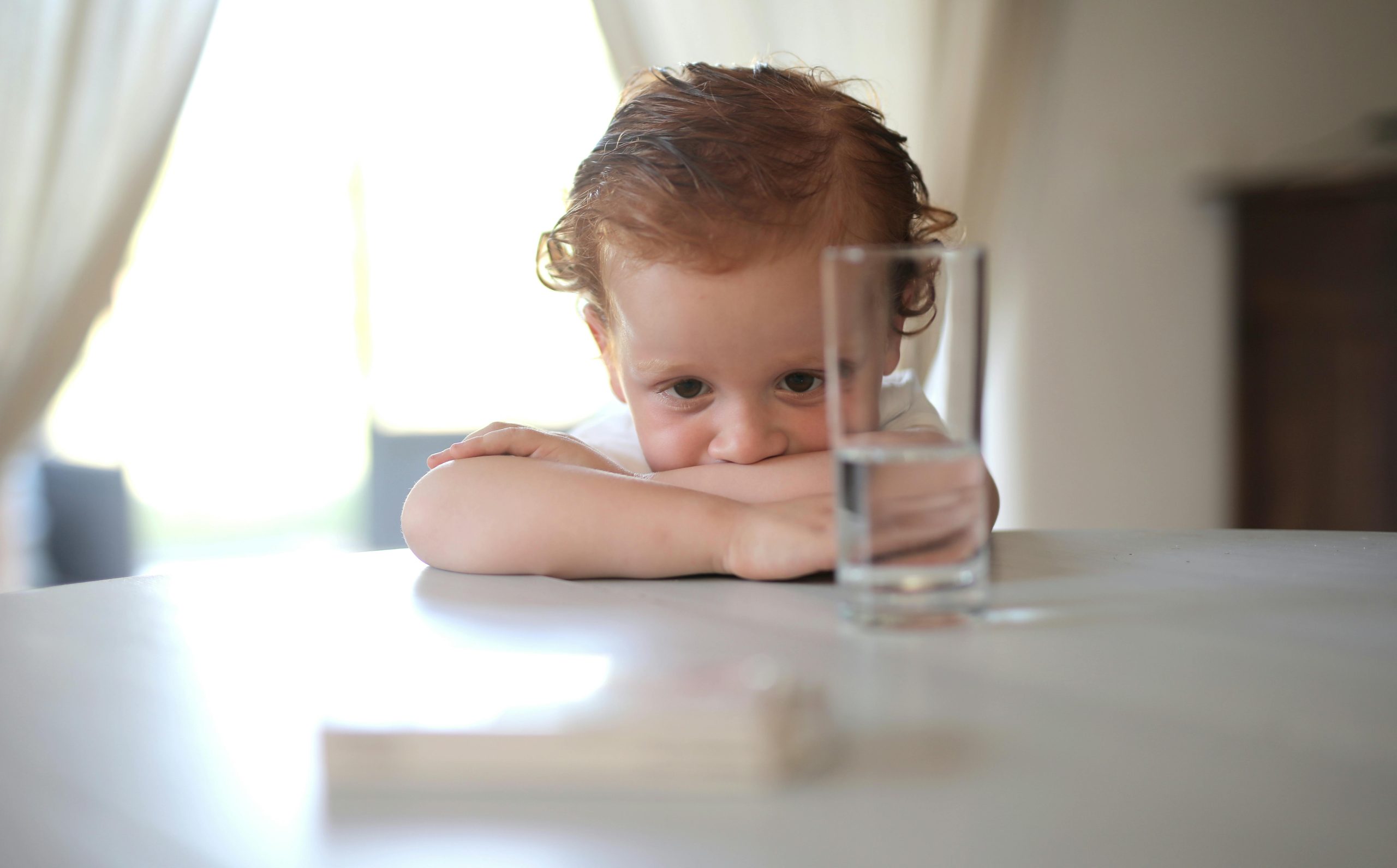 A cute child leans on a table indoors, gazing through a glass of water. Bright and serene setting.