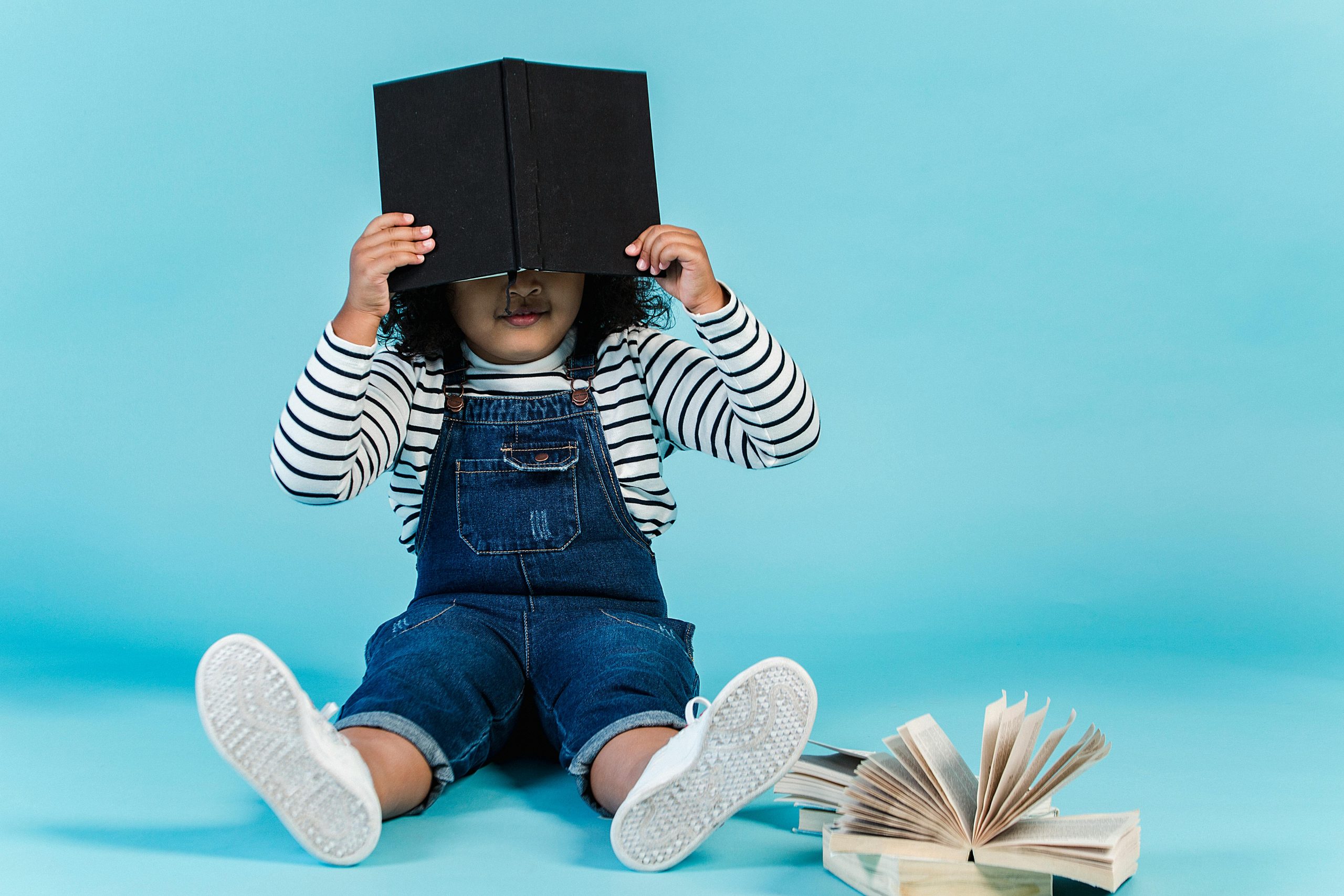 Unrecognizable little African American girl in trendy clothes sitting on floor with textbooks and covering face with book against blue background