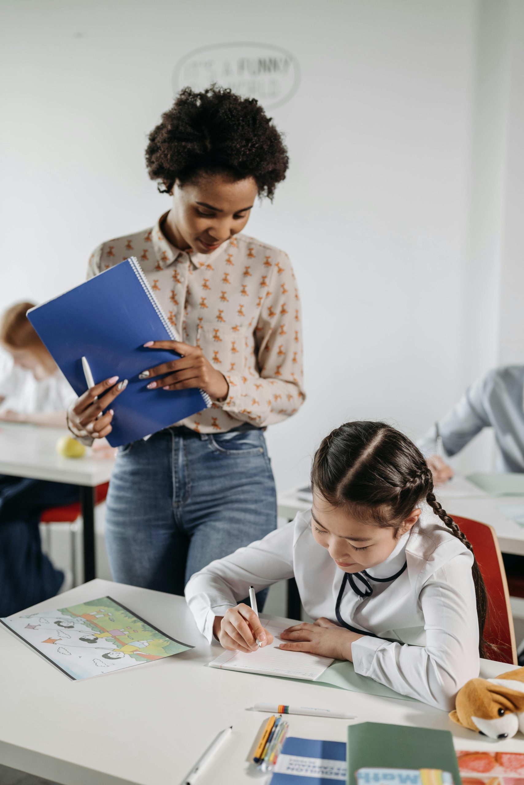 A diverse teacher helps a student with schoolwork in a classroom environment.