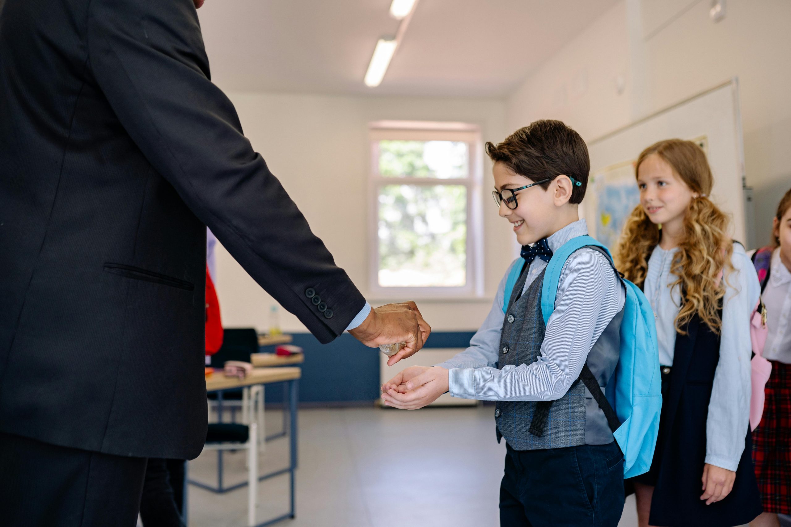 Children receiving hand sanitizer from a teacher in a classroom setting.