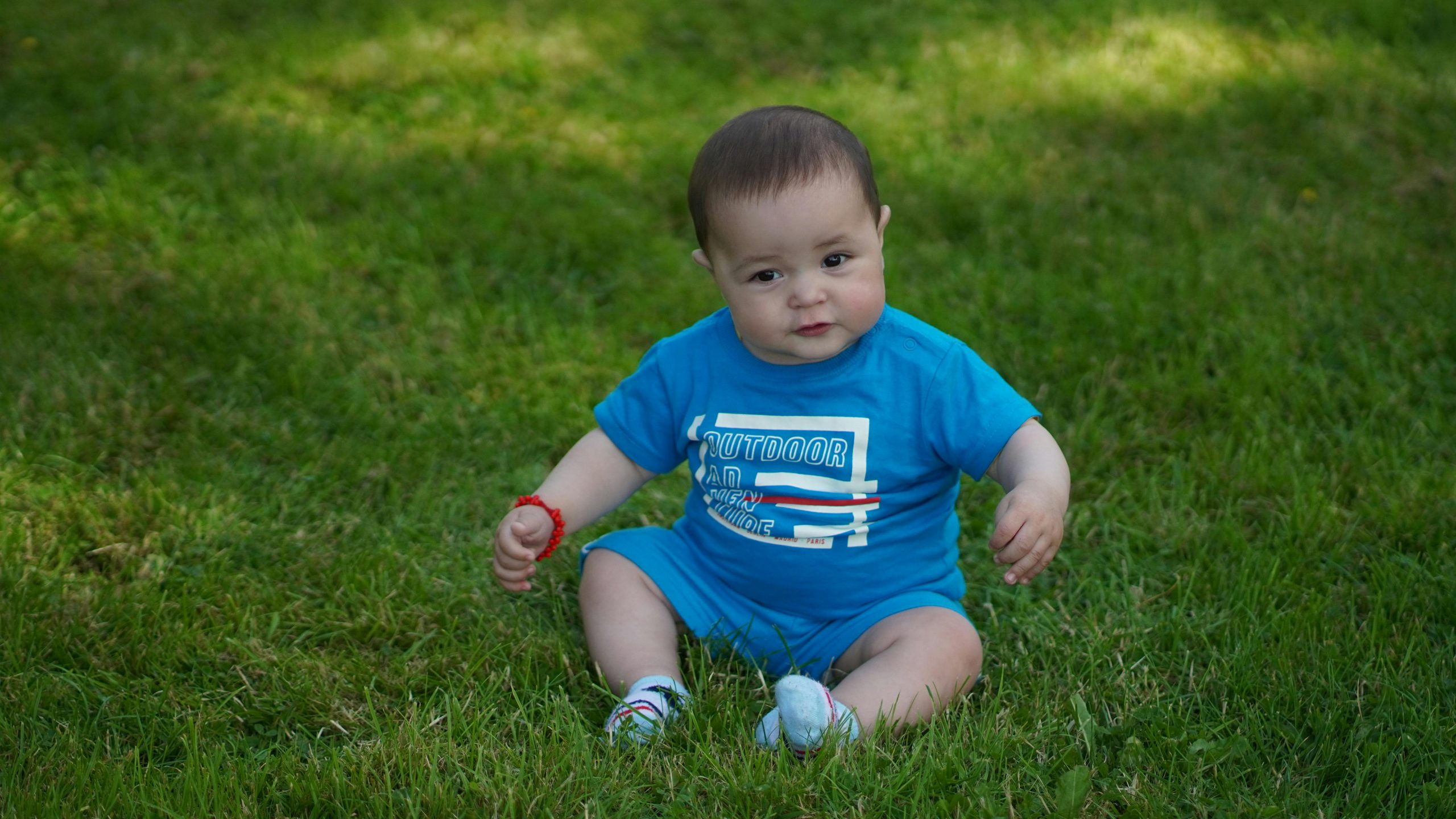 A cute baby in blue attire sitting on green grass during the day.